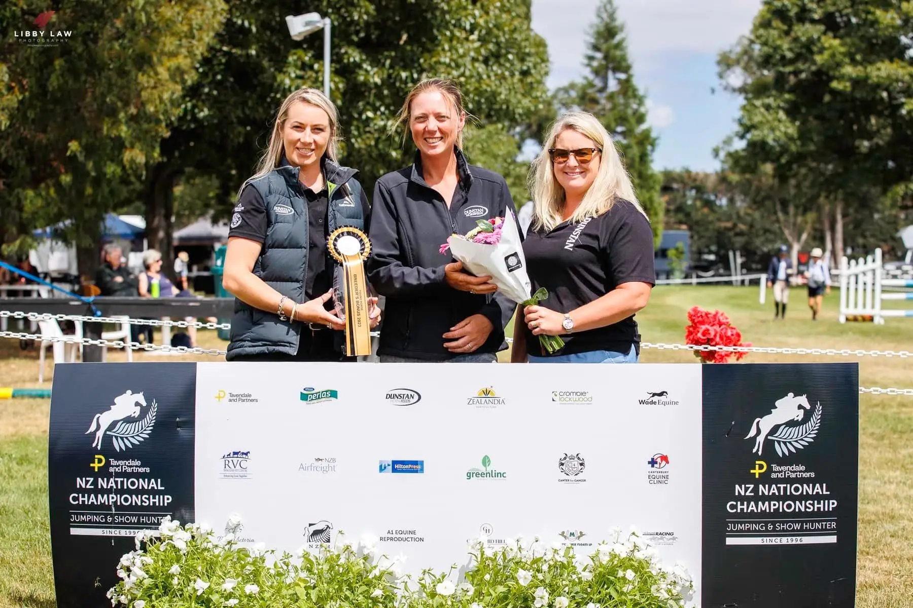 Three people with a trophy, rosette and flowers in front of the NZ National Championship Jumping and Show Hunter sponsor board