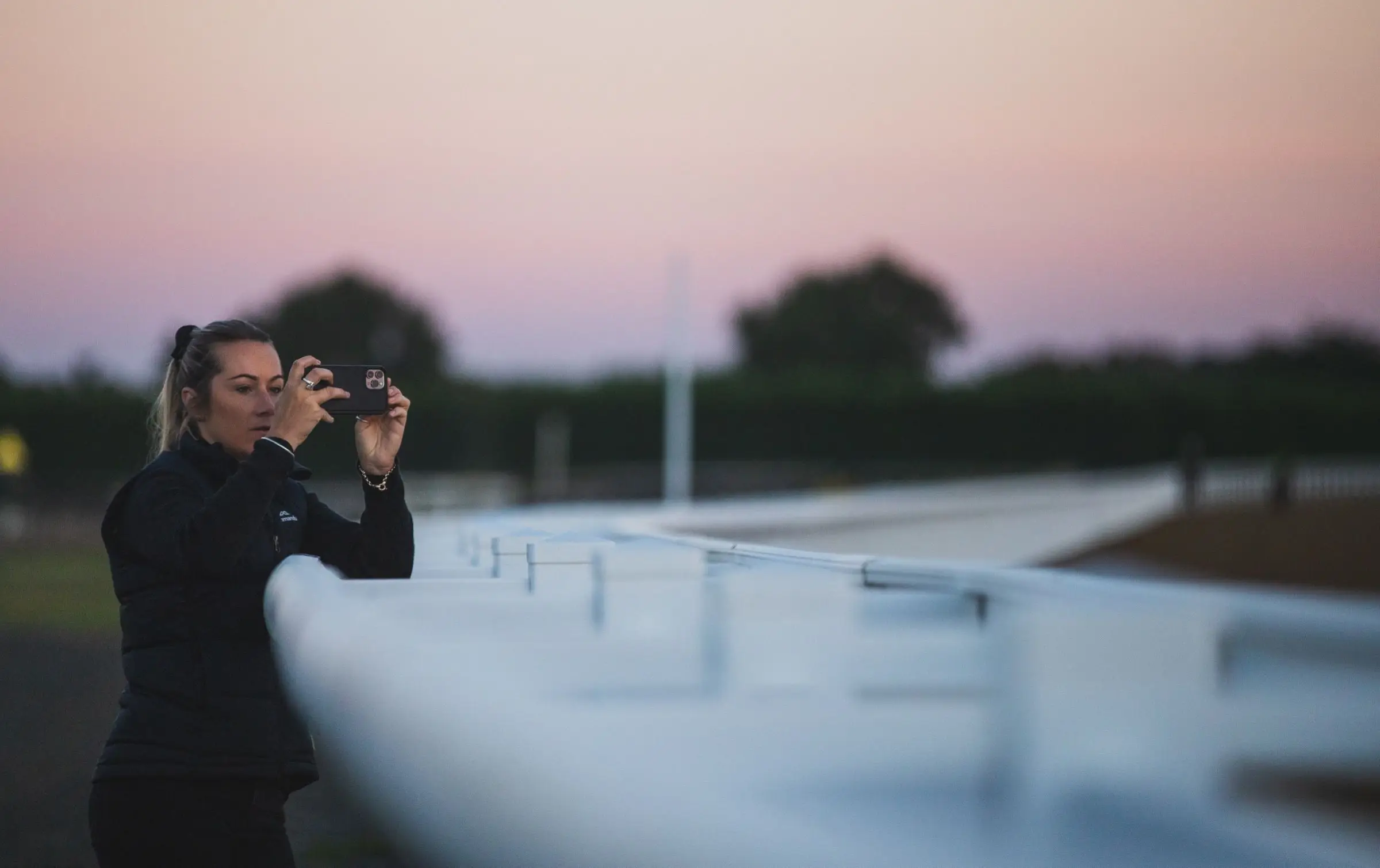 A woman filming on a phone over a white running rail at a raceway, pink dusk sky behind