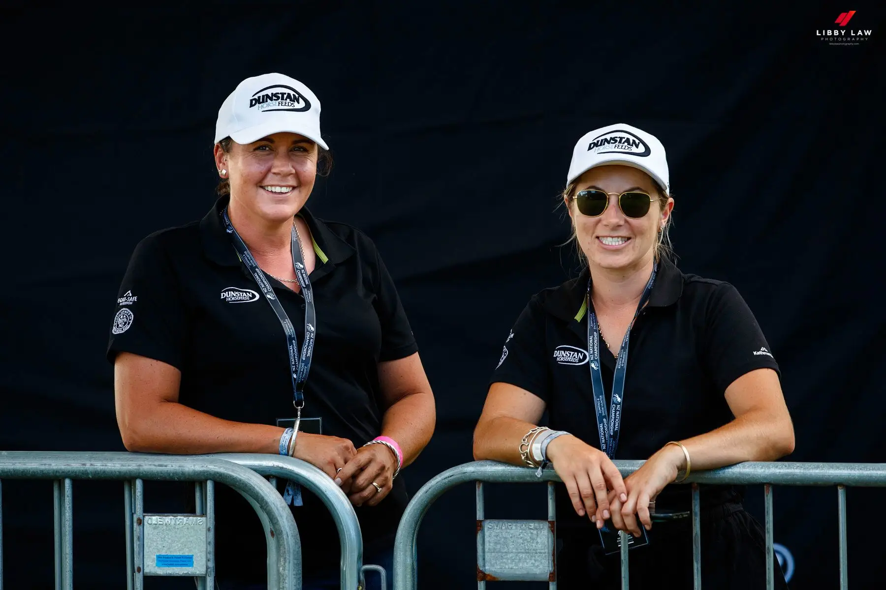 Two women in Dunstan Horsefeeds caps and polos leaning on a barrier at a show