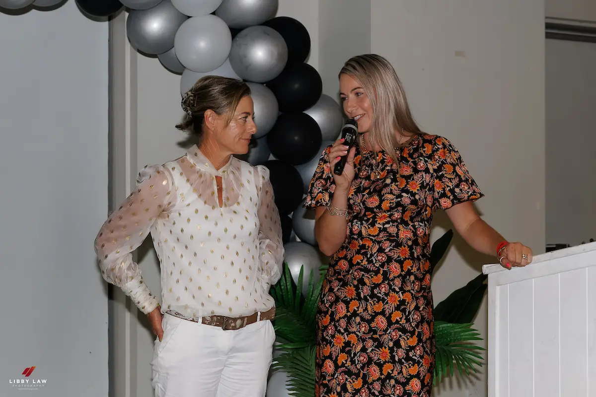 Paxton speaking into a microphone beside a guest at an awards dinner in front of a balloon arch