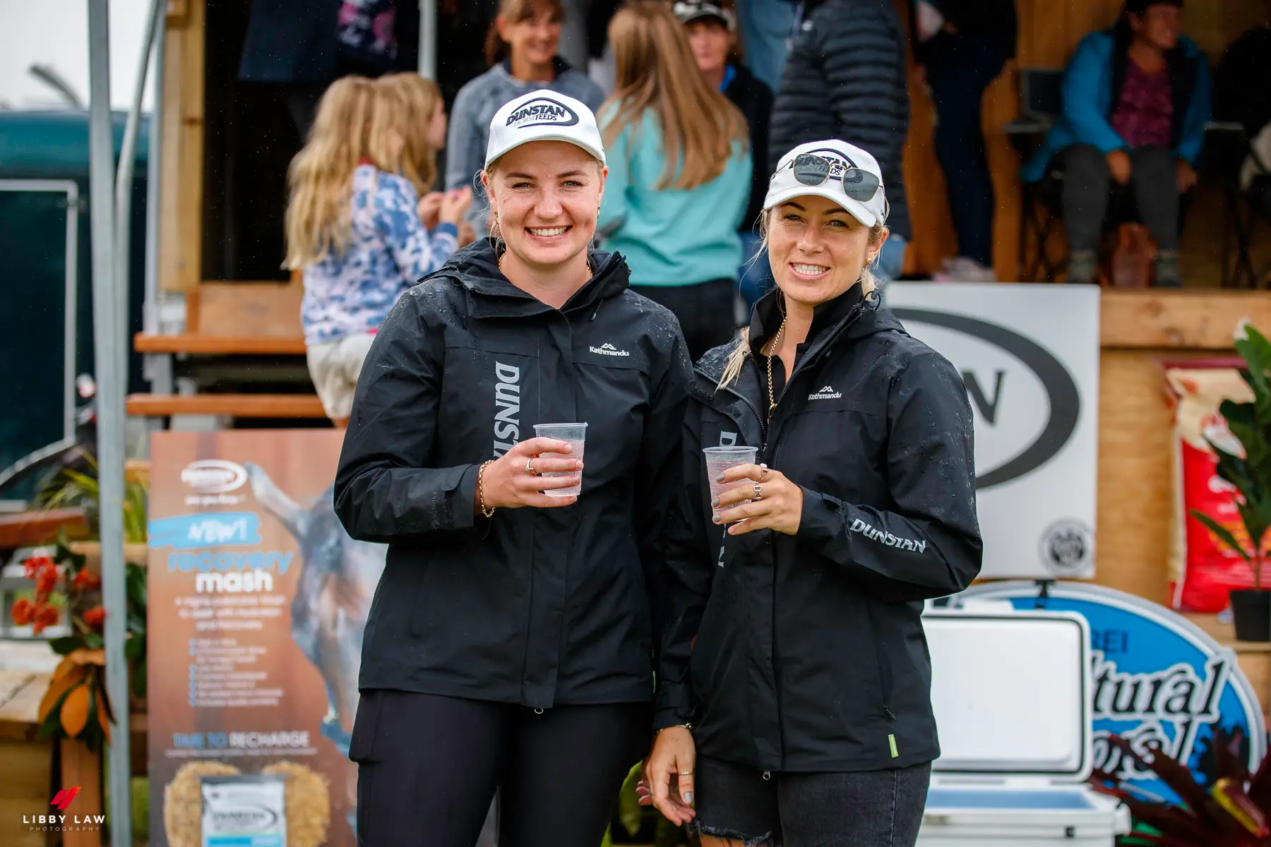 Two women in Dunstan Horsefeeds jackets and caps holding drinks at a wet event site