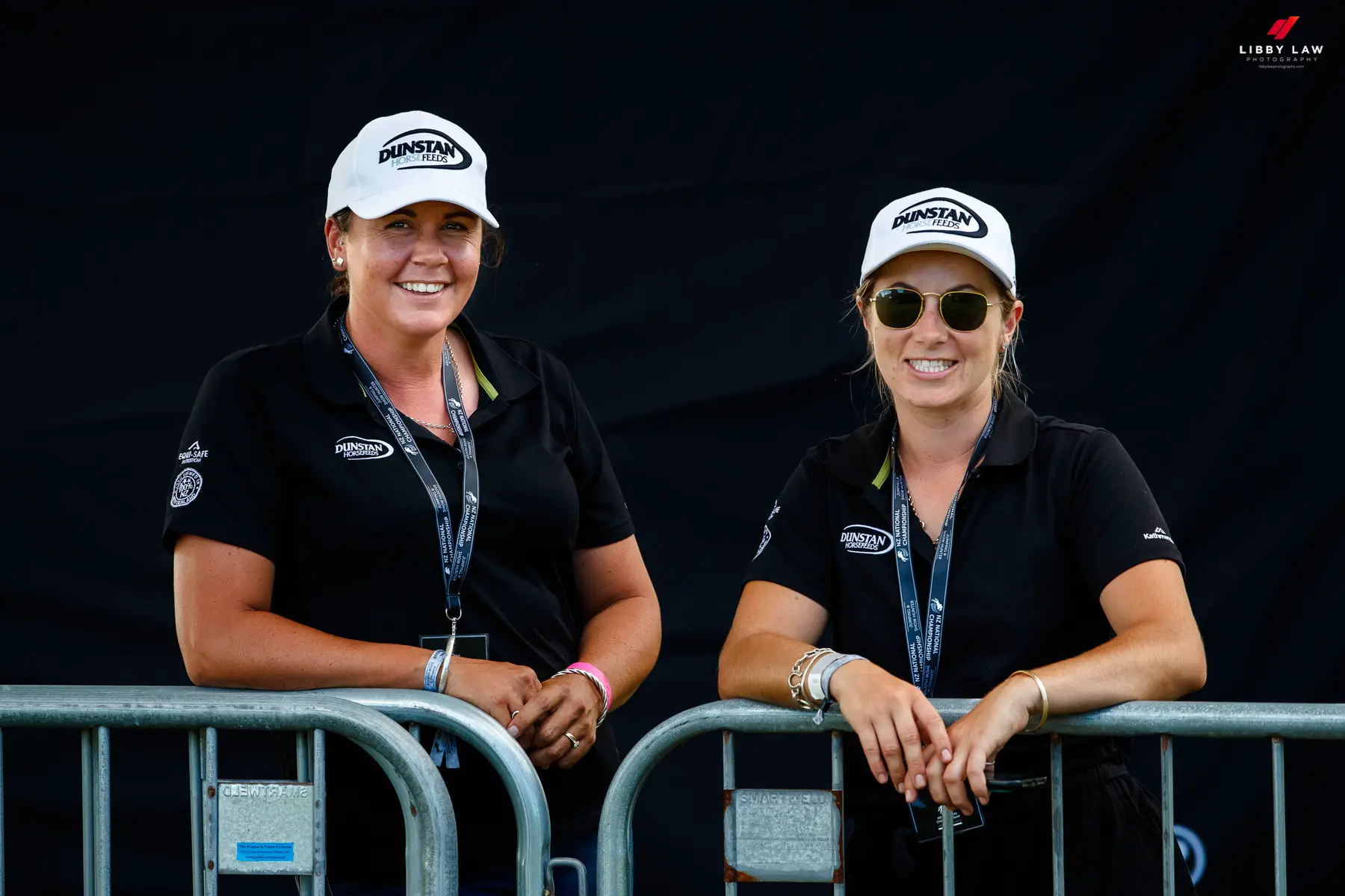 Two women in Dunstan Horsefeeds caps and polos leaning on a barrier at a show