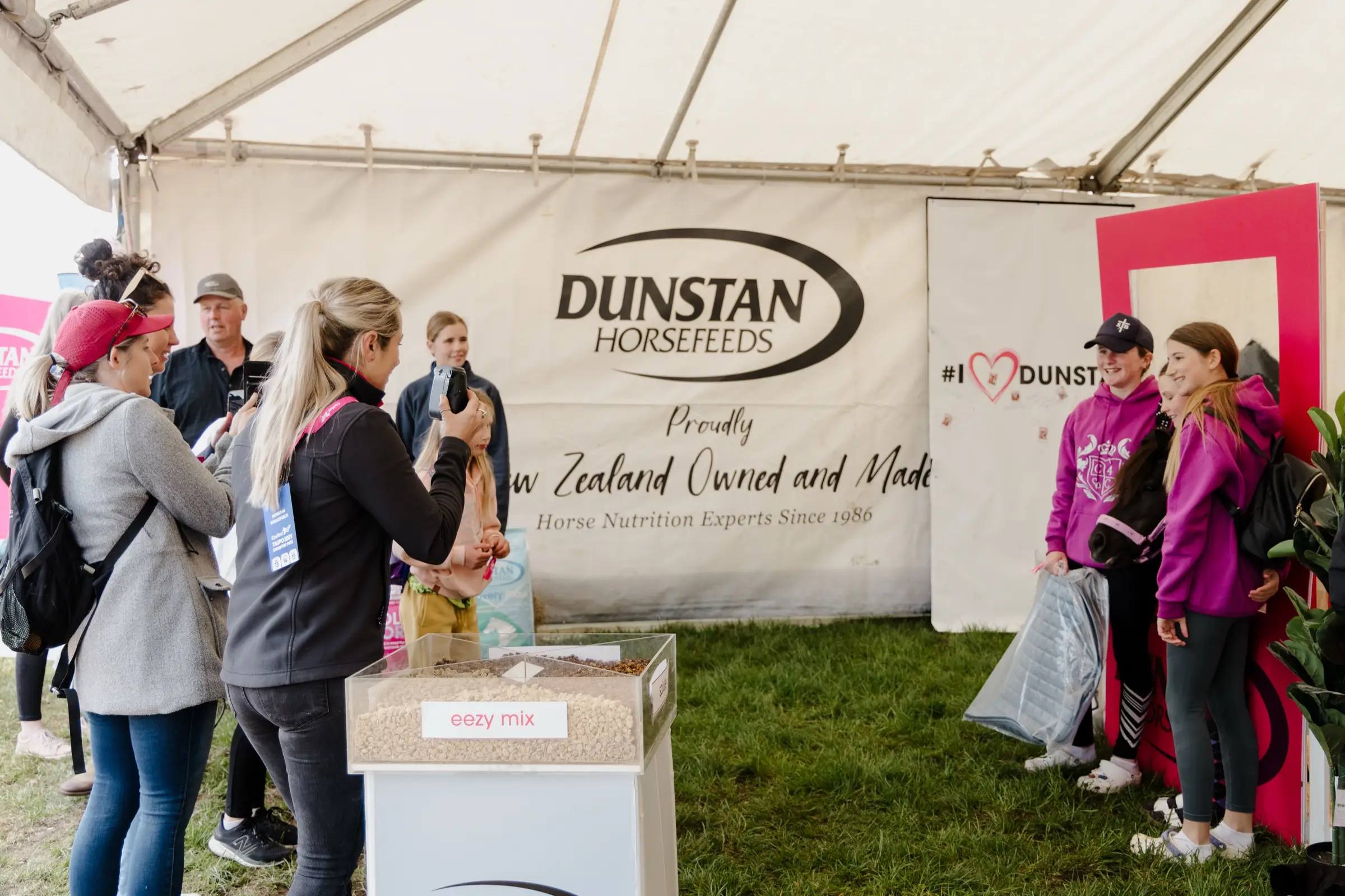 Visitors at a Dunstan Horsefeeds marquee at an event, a feed display in front and a branded photo frame to one side