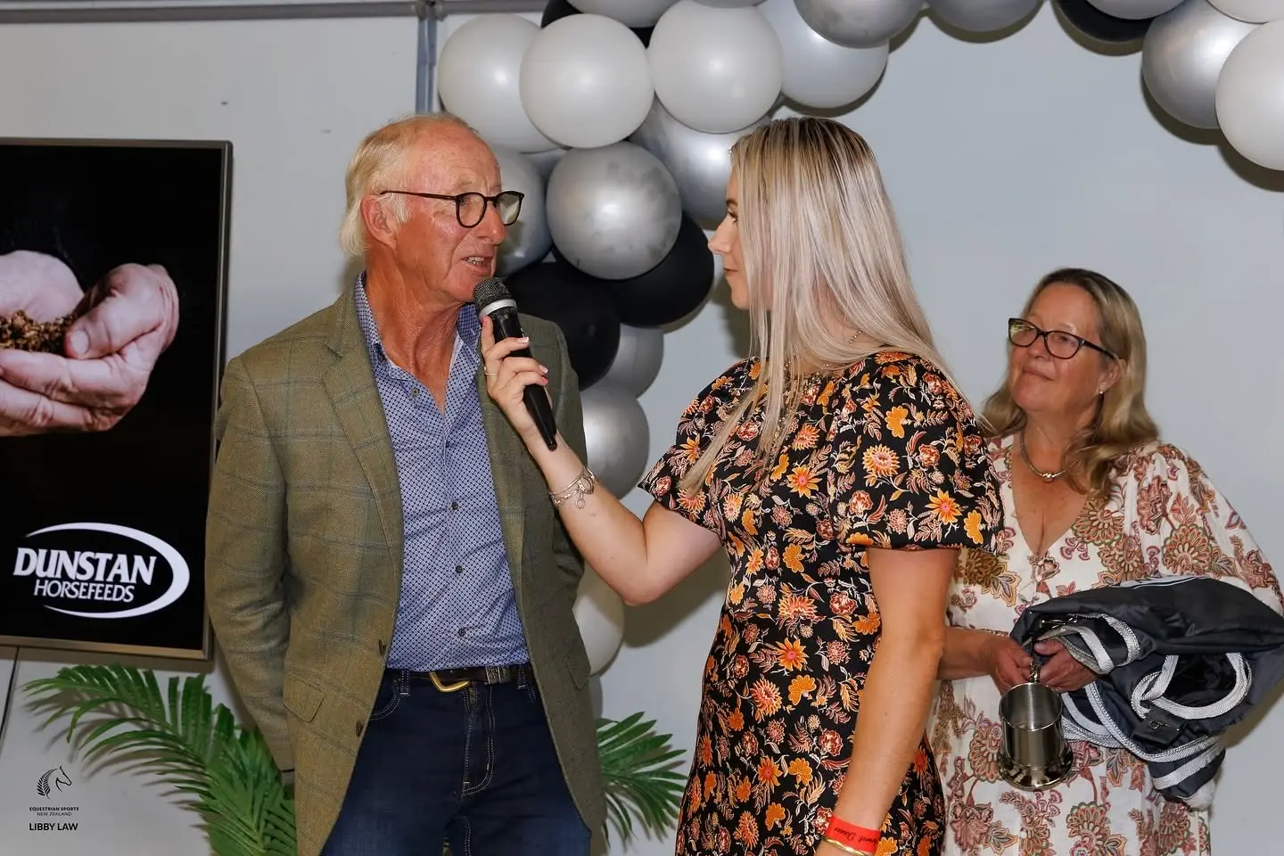 Paxton holding a microphone and interviewing a man at a Dunstan Horsefeeds function in front of a balloon arch
