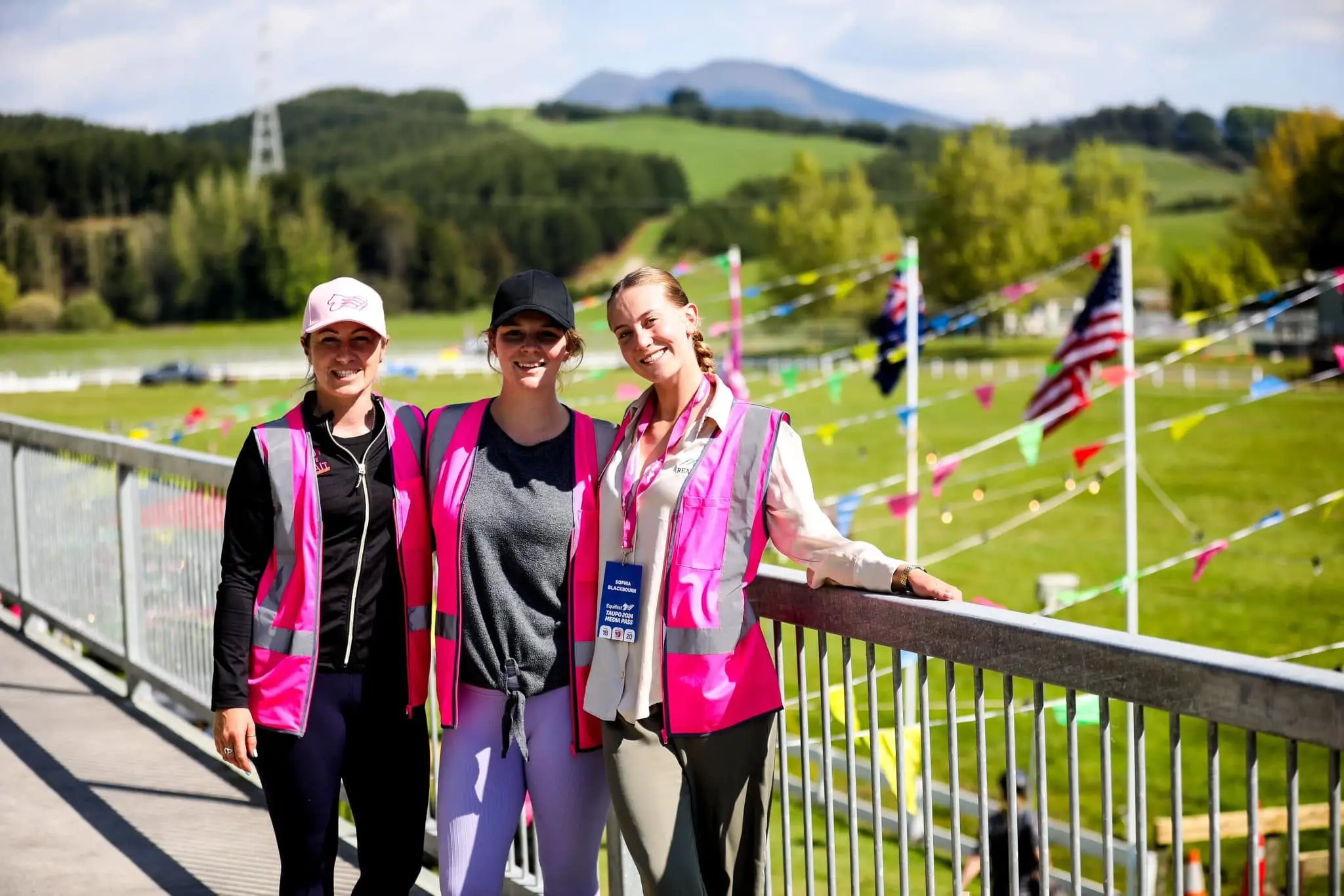Three event crew in magenta high visibility vests on a bridge above an event ground strung with bunting