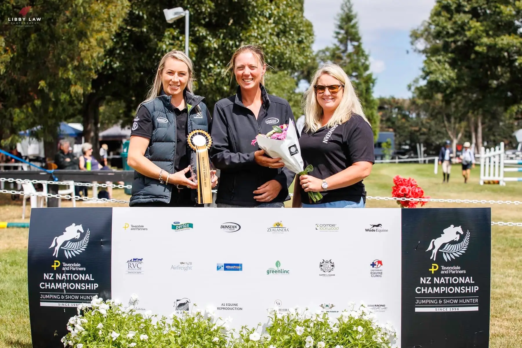 Three people with a trophy, rosette and flowers in front of the NZ National Championship Jumping and Show Hunter sponsor board