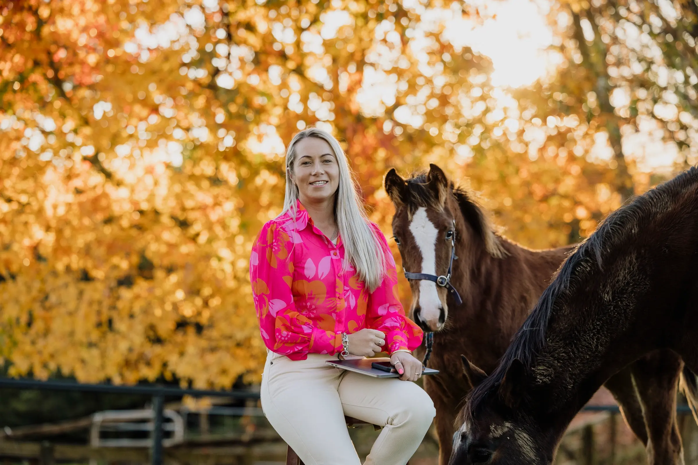 Paxton sitting on a paddock rail in a pink and orange floral shirt, laughing, with two horses beside her under autumn trees