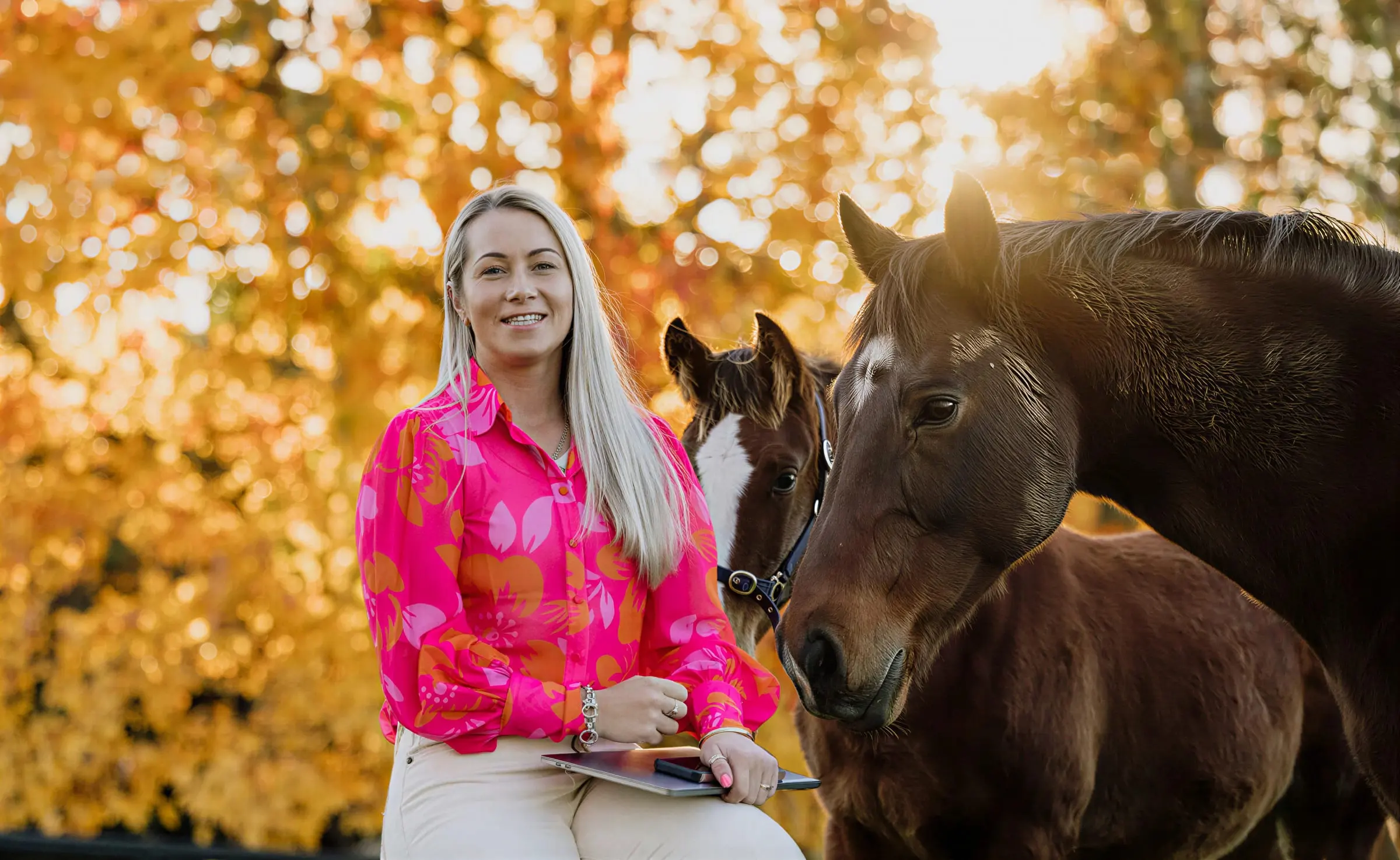 Paxton in a pink and orange floral shirt sitting with a tablet on her knee, a bay horse nosing in close beside her and a second horse behind, autumn trees lit low behind them
