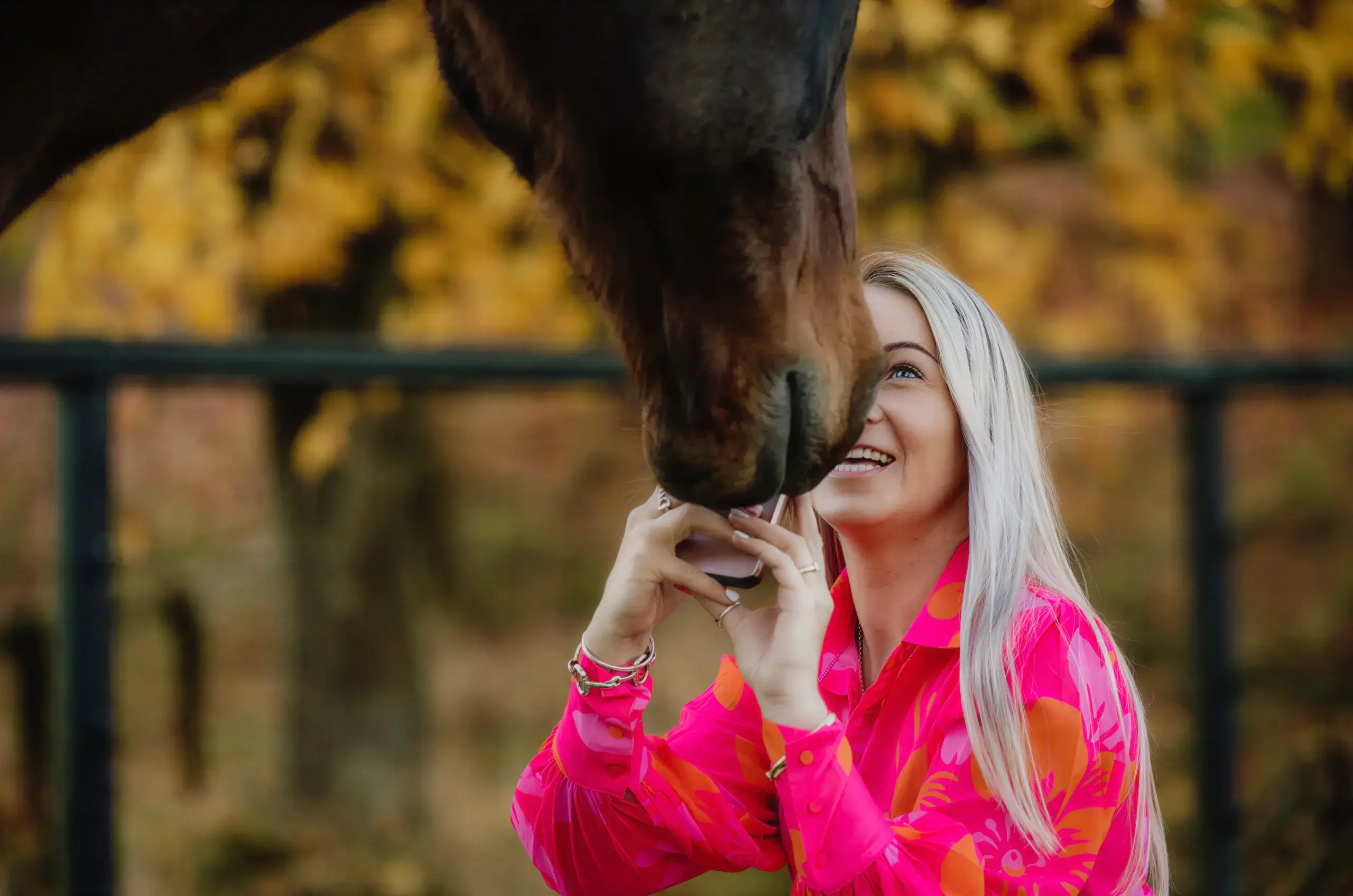 Paxton laughing up at a bay horse nosing her phone, autumn leaves behind