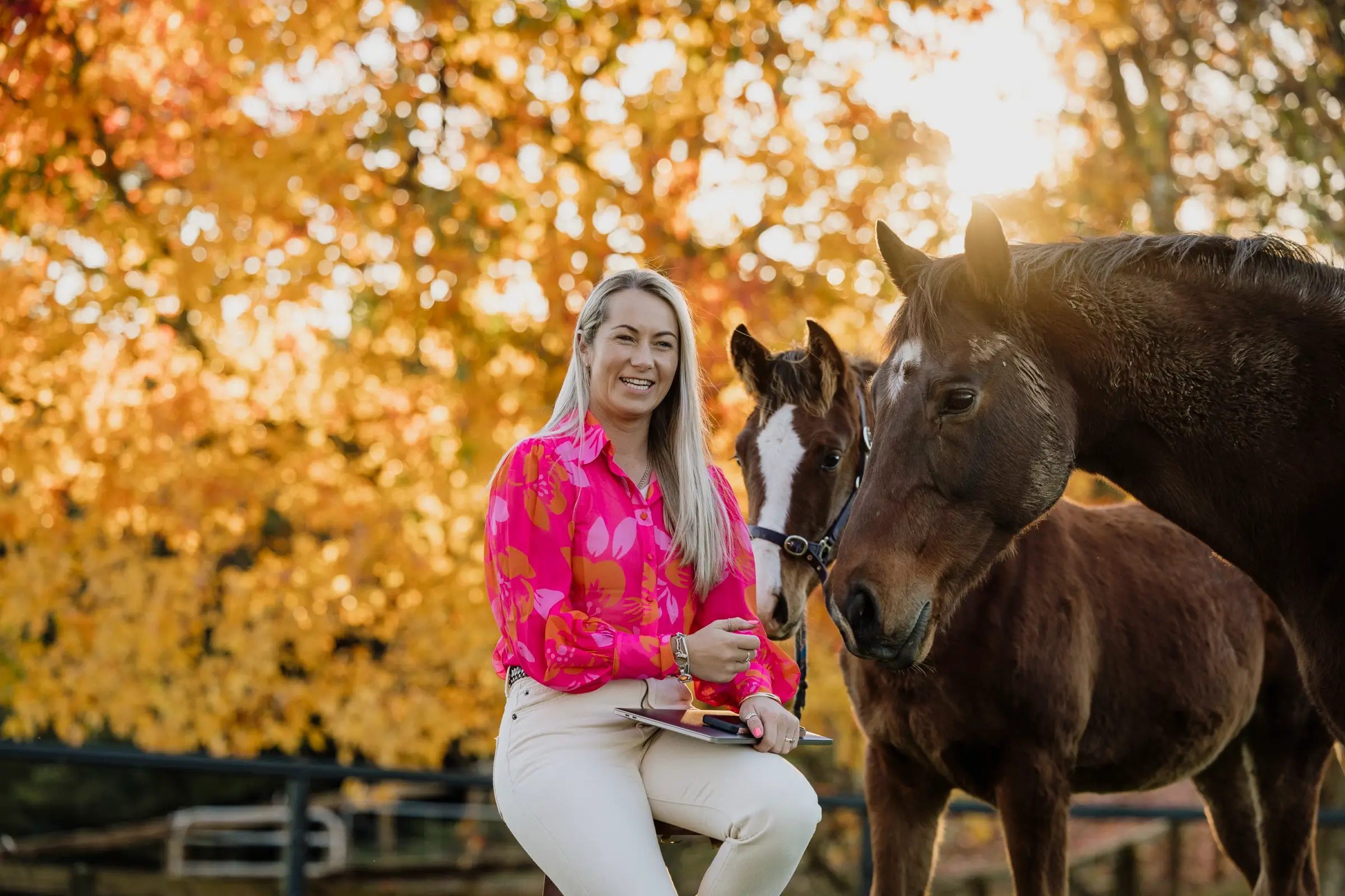 Paxton laughing on a fence rail while two horses lean in towards her, golden autumn leaves behind
