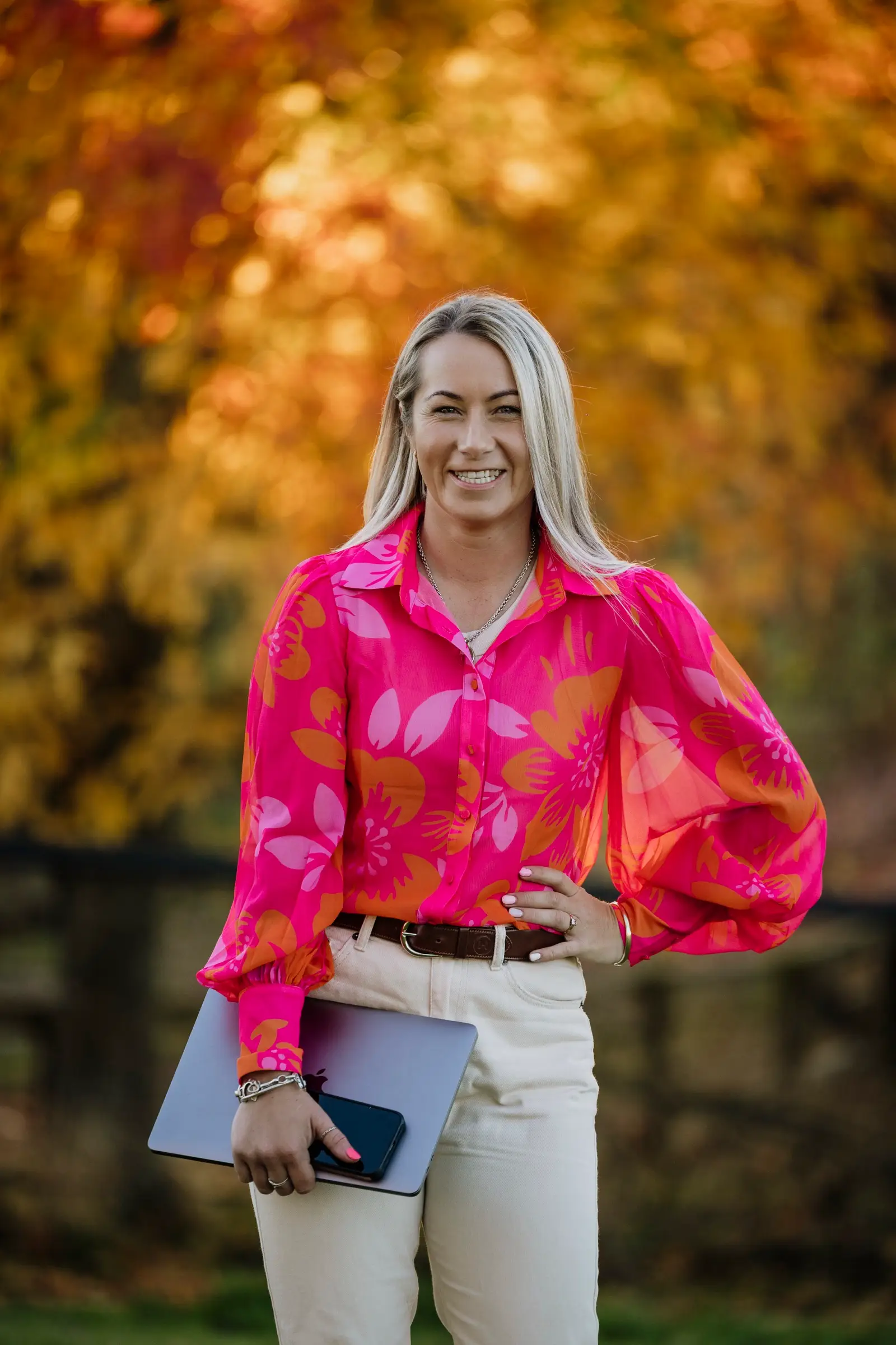 Portrait of Paxton standing with a laptop and phone under one arm, hand on hip, autumn colour blurred behind her