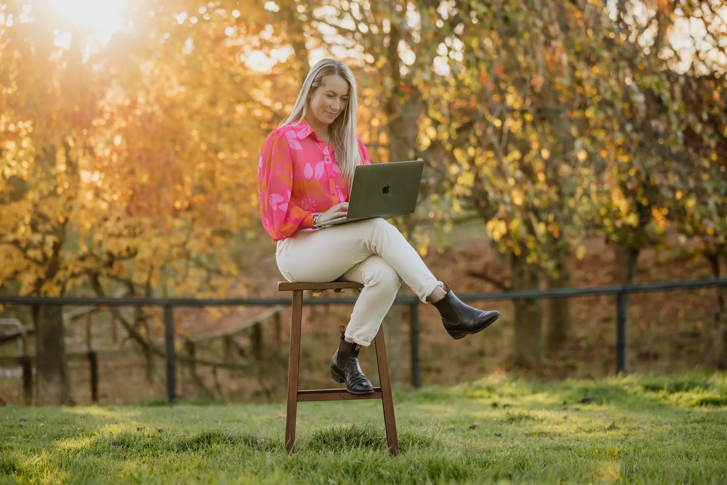 Paxton perched on a wooden stool in the middle of a paddock with a laptop open on her knee, backlit by low sun
