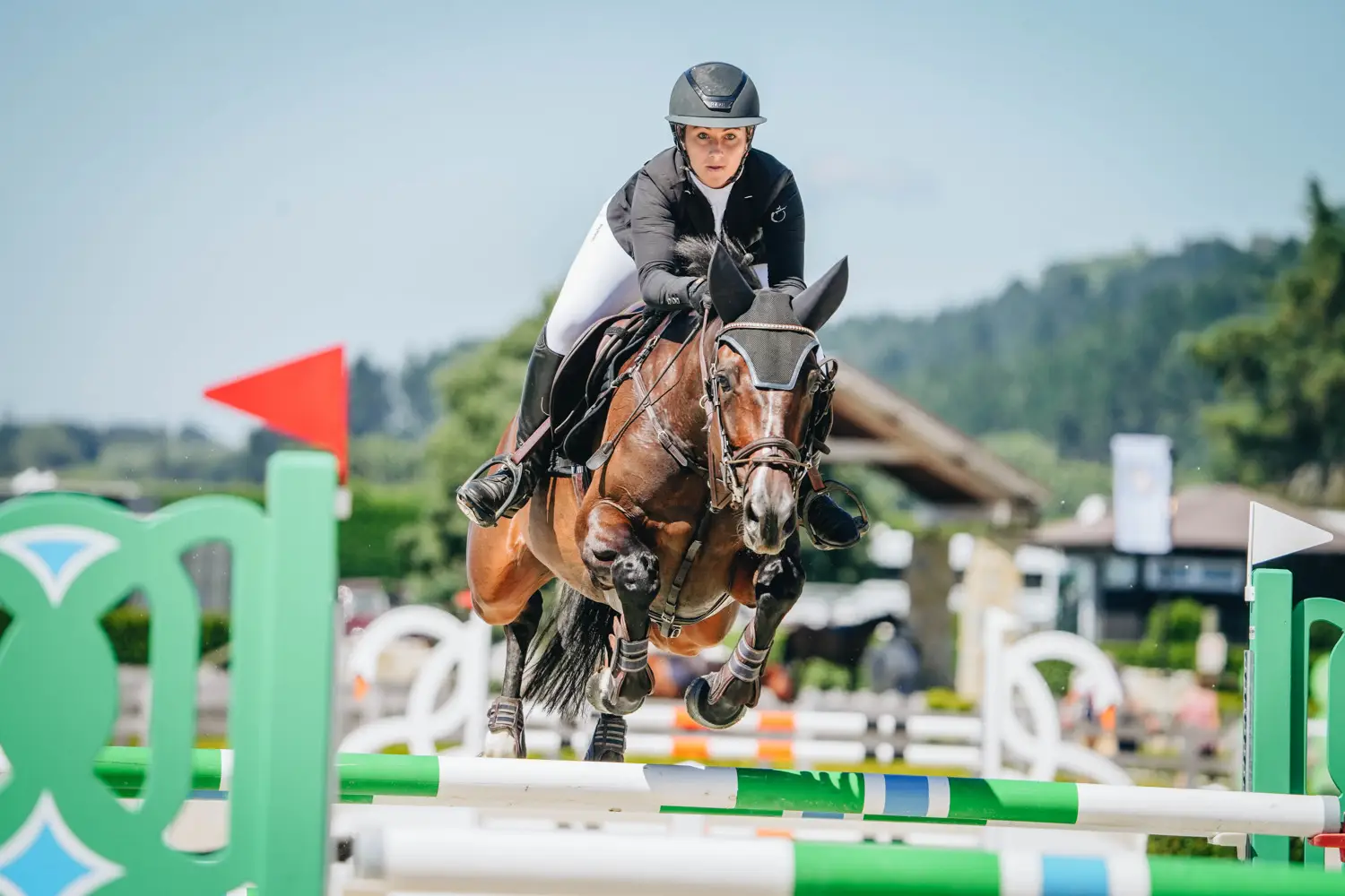 A horse and rider jumping a green oxer on a clear day with hills behind