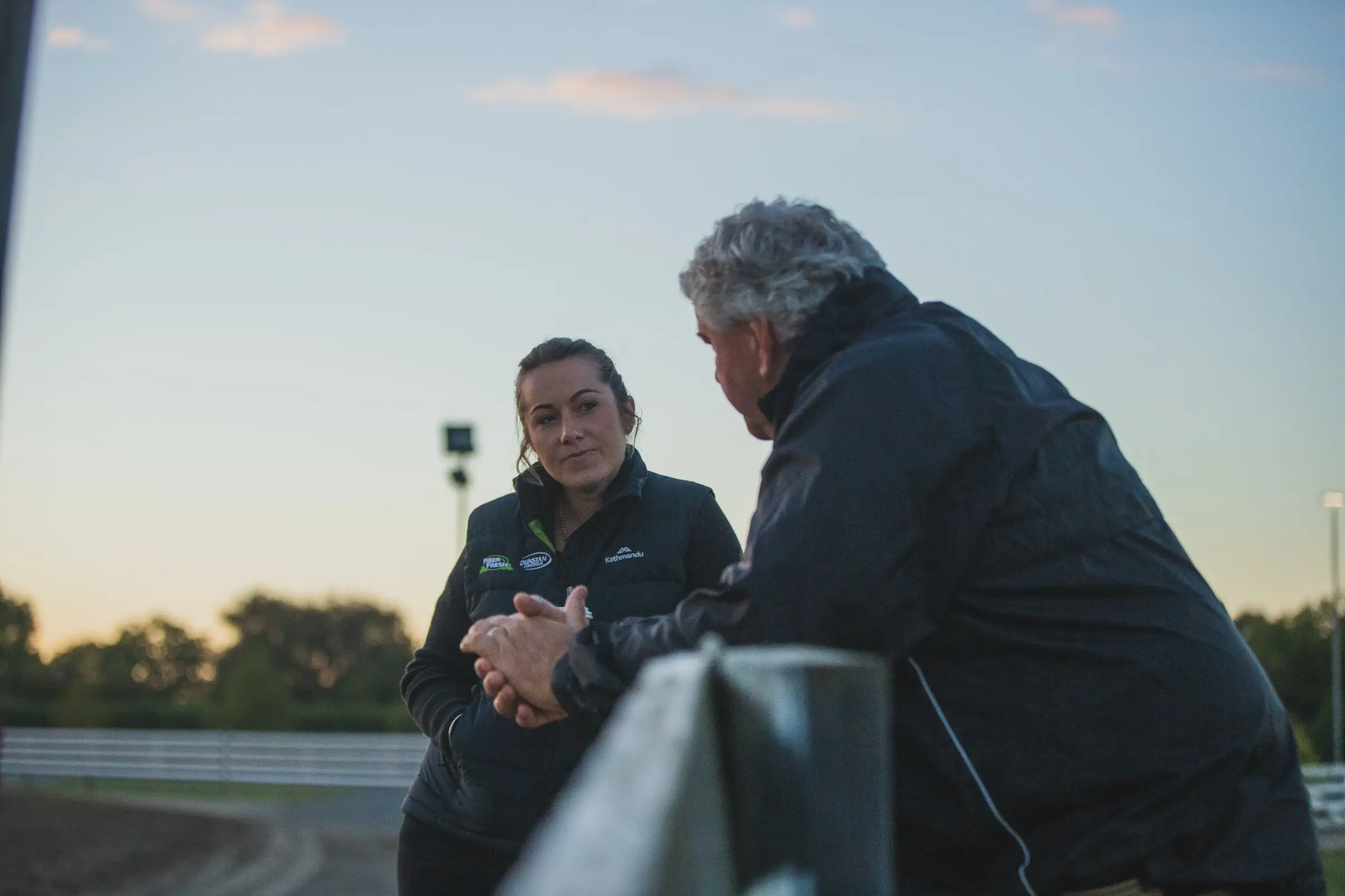 Two people in branded jackets talking across a rail at a raceway at dusk