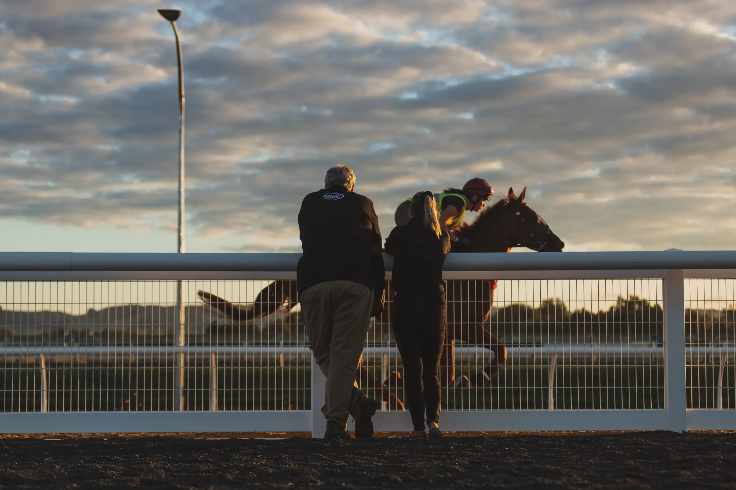 Two people watching from behind a white running rail as a horse and rider work at dawn under a wide cloudy sky