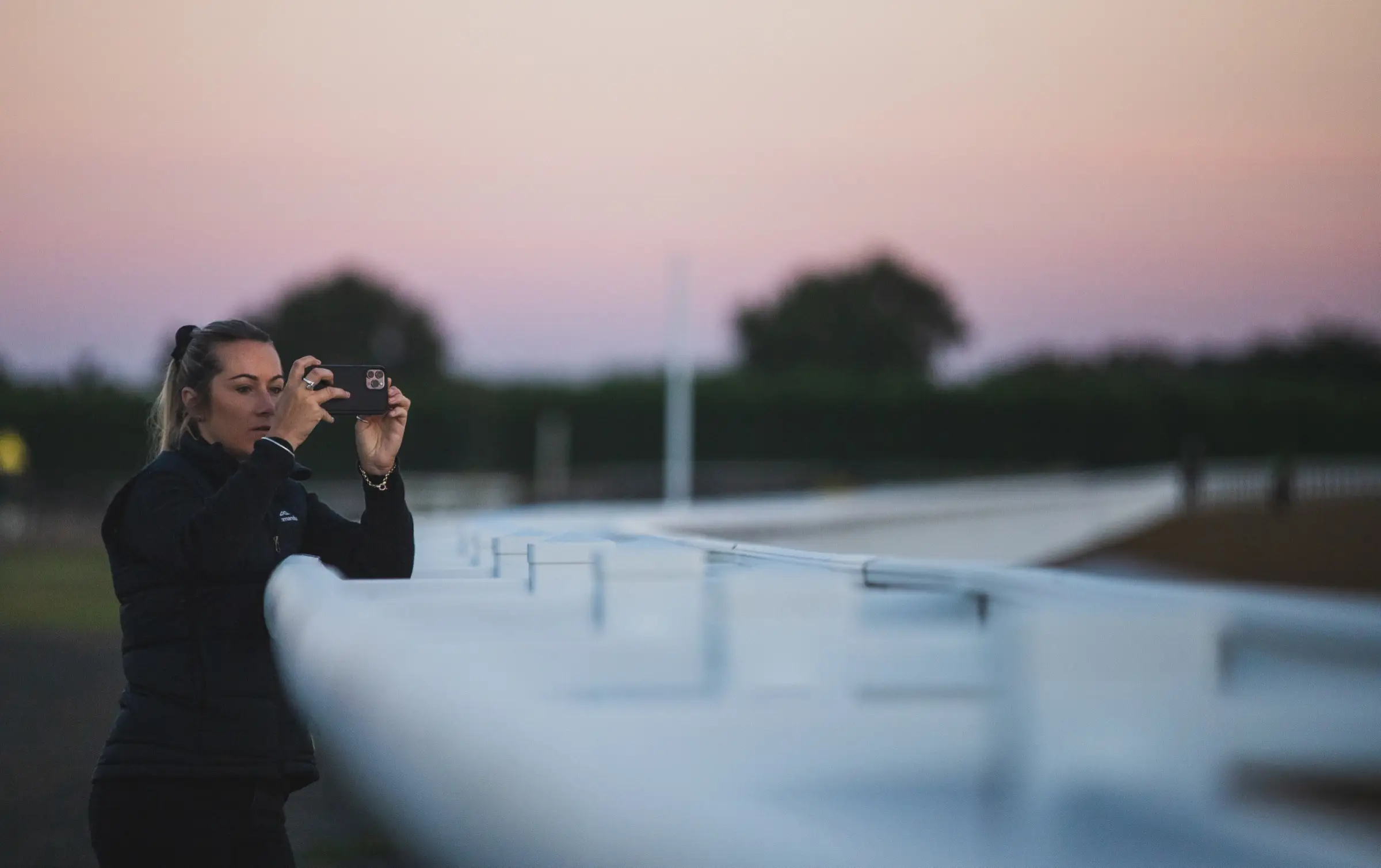 A woman filming on a phone over a white running rail at a raceway, pink dusk sky behind