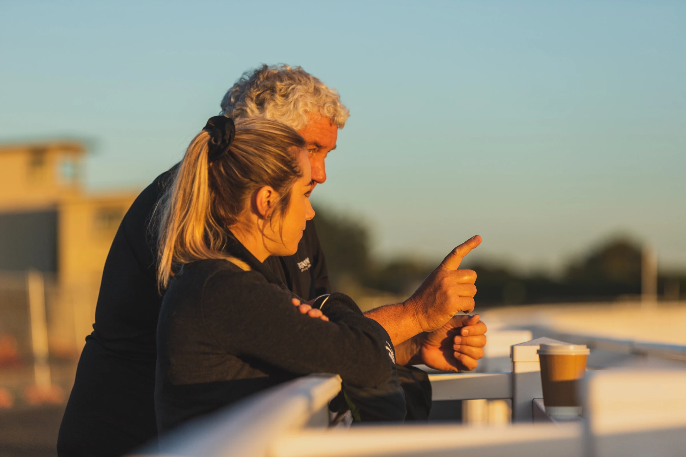 Two people leaning on a rail in low golden light, one pointing down the track, a coffee cup beside them