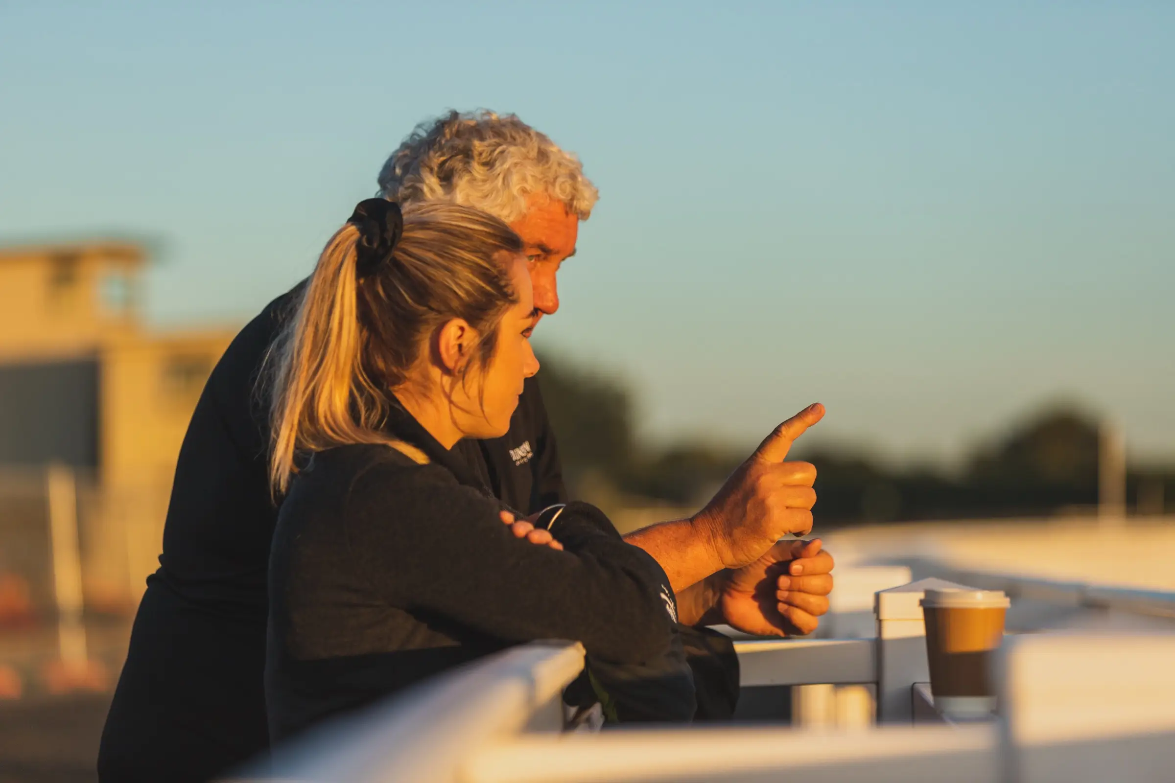Two people leaning on a rail in low golden light, one pointing down the track, a coffee cup beside them